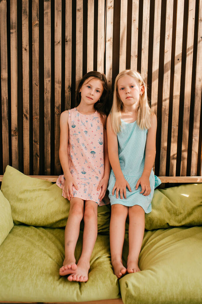 Two little model girls in beautiful dresses sitting on lemon bed with wooden wall on background and posing for camera - Foto, Bild