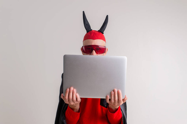 Stylish caucasian man in devil hat with horns and vampire cape with laptop isolated on white backhround. - Foto, afbeelding