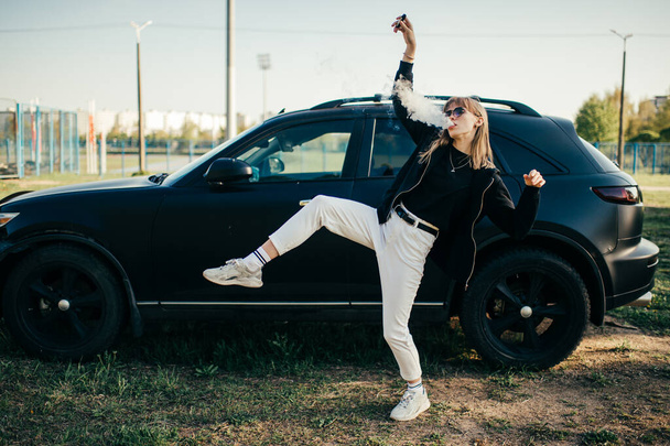 Bella giovane donna caucasica in jeans bianchi, scarpe da ginnastica bianche e giacca nera si trova vicino alla vecchia auto nera fuori. - Foto, immagini
