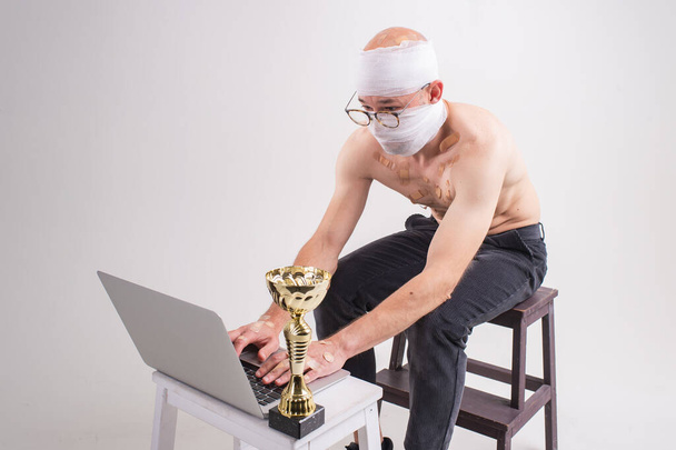 Injured man with many patches on his body and bandaged head shows his gold cup and serfing internet with laptop isolated on white background - Фото, зображення