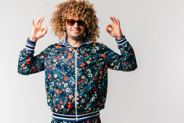 Adult positive smiling funky man with afro curly hair style in suglasses, multicolored clothes making okay gesture with hands on white studio background. Funny portrait of artistic stylish male person - Photo, Image