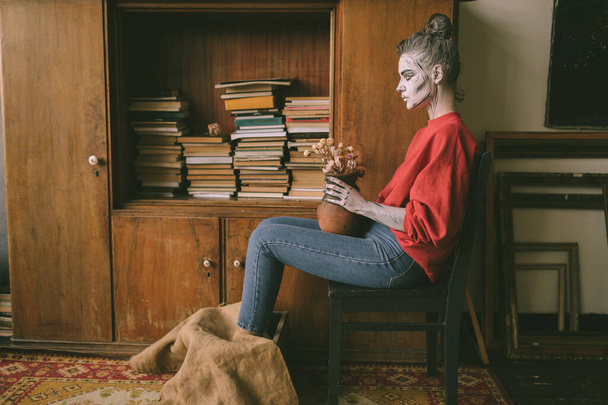 Dziwna kobieta wirh Conoc pasek ciało sztuka siedzi na krześle z wazonem suchych kwiatów w rękach przeciwko szafie z książkami. - Zdjęcie, obraz