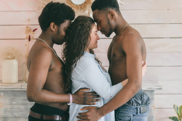 Multicultural love and relationships concept. White sexy girl embracing two topless dark skinned men. Indoor odd portrait of interracial loving trios. Two passionate african men hugs their girlfrined - 写真・画像