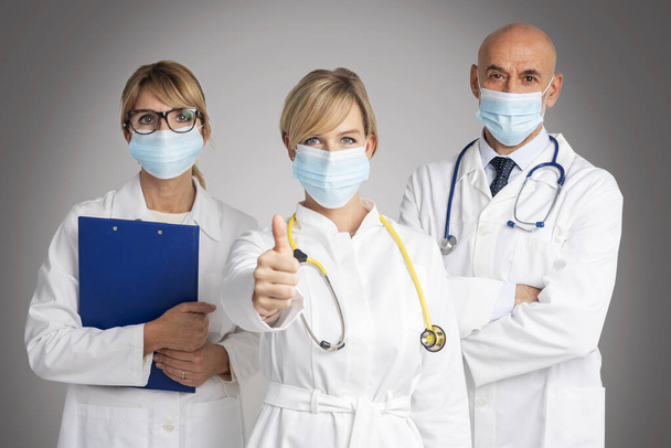 Shot of female and male doctors standing together at isolated grey background while female doctor giving thumbs up. Medical team. Doctors wearing face masks for prevention. - Foto, imagen