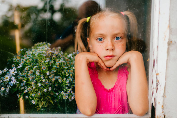 Little cute caucasian self isolated girl with long fair hair and in pink dress holds flowers and smiles behind the window  during pandemia of coronavirus covid 19.  Stay home. Social disntancing. - Foto, immagini