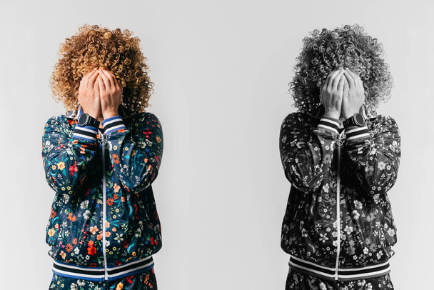 Black and white and color portrait of unrecognizble unhappy depressed man with face hidden by hands standing on white background beyond his alter ego. Self reflection. Afro male in tracksuit crying - Фото, изображение