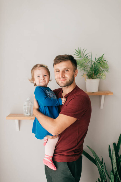 Caucasian handsome strong dad in a bright room hold his cute little daughter which sits on a shelf - Foto, Imagen