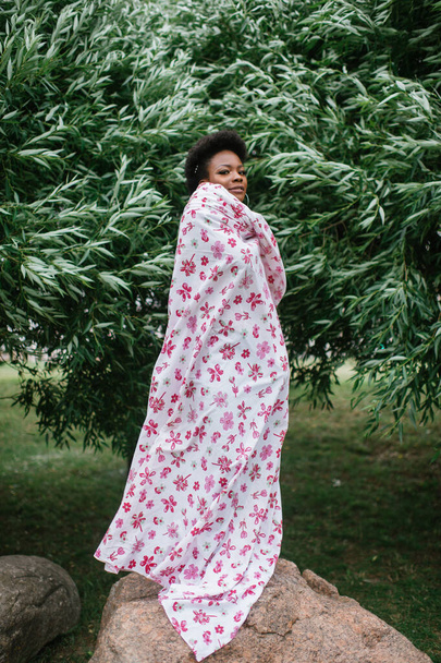 Beautiful dark skinned girl wrapped in sheet with fashion print posing at park with trees on background. - Foto, Bild