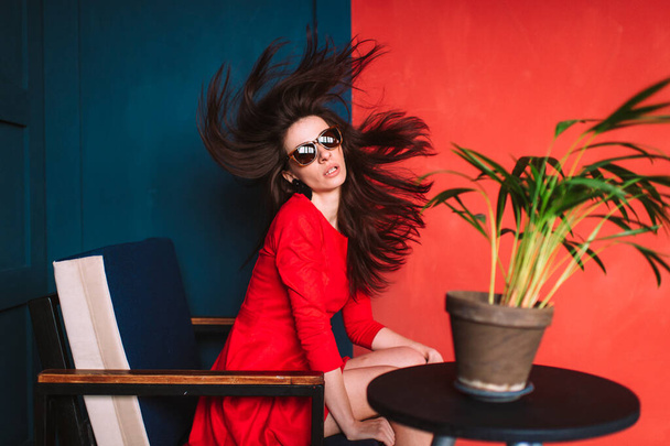 Beautiful fashion girl with black long hair, in sunglasses, red elegant dress sitting in chair on blue red wall in studio. Stylish brunnetre with scattered flying hair. Hairdresser services concept - Fotografie, Obrázek