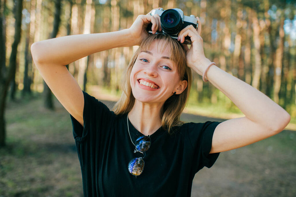 Portrait of beautiful young female photographer with short fair hair in black t-shirt stands with her camera and smiles. - Foto, imagen