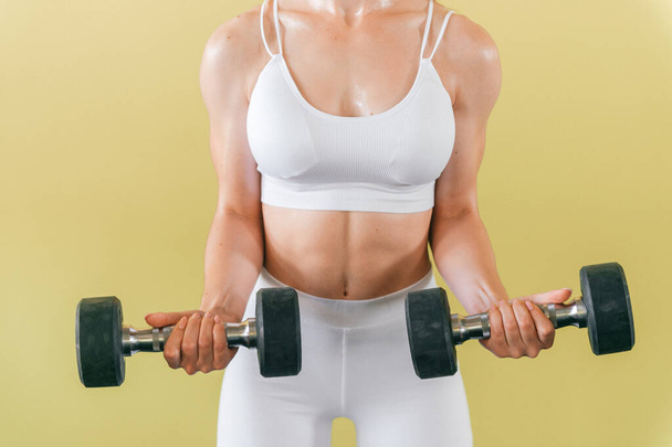 Atletische bodybuilder vrouw met halters. Mooi brunette meisje met spieren tillen gewichten op gele achtergrond - Foto, afbeelding