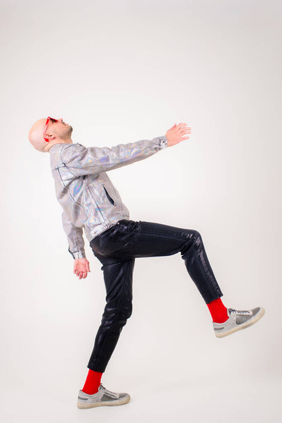 Strange caucasian man without hair in grey shirt, black leather trousers, black sunglasses, red socks and grey trainers rejoices isolated on white background - 写真・画像