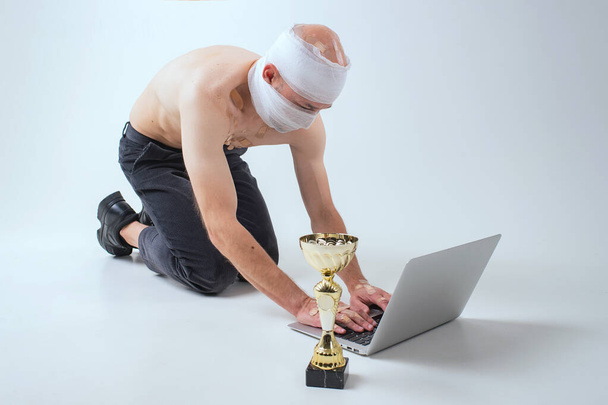Injured man with many patches on his body and bandaged head shows his gold cup and serfing internet with laptop isolated on white background - Photo, Image