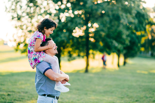 Dad carrying his daughter on shoulders in summer park at sunset. Father have fun with his little child. Kid siiting at parent neck. Happy family playing and making  crazy faces.  Lifestely portrait. - Fotografie, Obrázek
