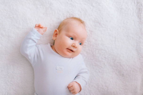 Adorable lovely newborn male baby with blue eyes lifestyle indoor portrait from above. Cute child with hands apart lying on back in pijama on bed at home. Little child with emotional face rest on sofa - Φωτογραφία, εικόνα