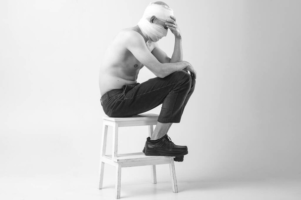 Injured man with many patches on his body and bandaged head sits on a chair and covers his hands on face isolated on white background - Foto, Imagem