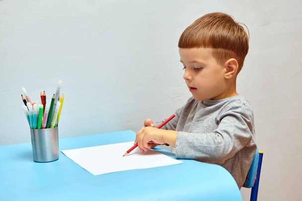 Mignon petit garçon aux cheveux blonds dessine des crayons de couleur à la maison. Dessine à la table bleue. - Photo, image