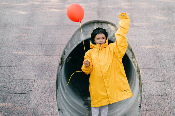 Portrait of pretty litle girl in big adult oversized yellow raincoat with red balloon in hand standing and sitting against sewage tunnel pipe. Horror movie concept. Funny child posing in summer day - Fotoğraf, Görsel