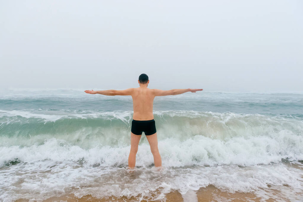 Odd bizarre crazy fearless awesome adult man enjoying in cold stormy sea. Guy jumping in waves in ocean with emotional funny face. Adult male fooling like child in water outdoors. Leisure activities - Foto, afbeelding