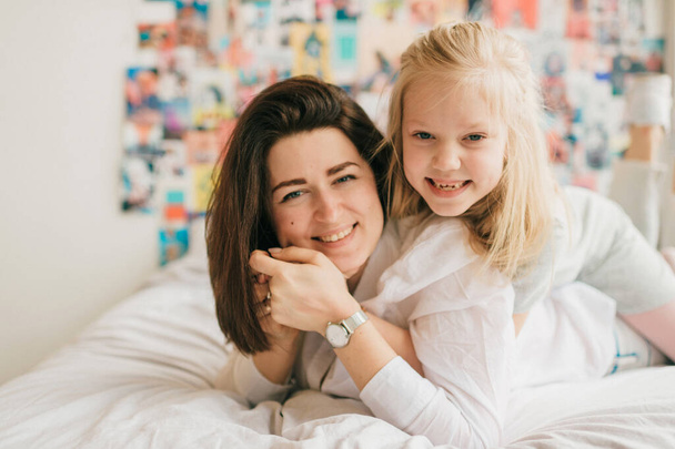 Lifestyle soft focus portrait of happy mom hugs her adorable daughter on white bed. Indoor happy family portrait of smiling mother and her daughter lying together on bed with decorative wall behind - Zdjęcie, obraz