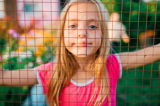Retrato de hermosa chica caucásica con el pelo largo y rubio y en vestido rosa sonríe de pie detrás de una valla de malla en la aldea - Foto, Imagen