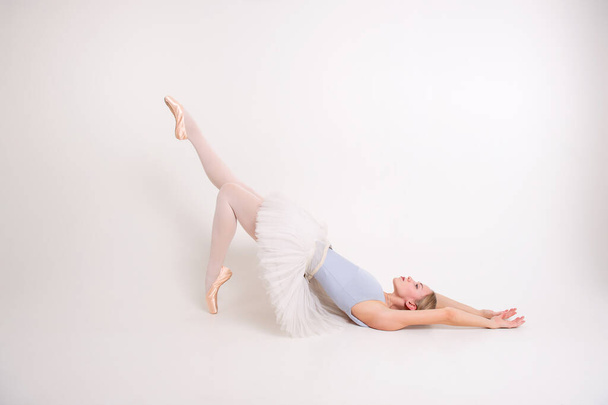 Attractive caucasian female with pale skin abd blonde hair in grey leotard, pointes and white skirt for ballet lies on the floor - Fotografie, Obrázek