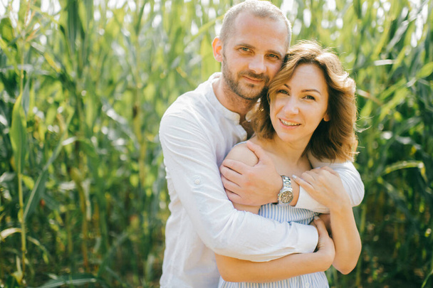 Attractive caucasian woman with short wavy dark hair in blue dress with her strong husband with short dark hair in white shirt and blue jeans relaxes together in the village cornfield - Foto, imagen