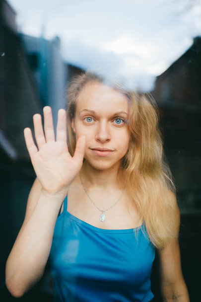 Portrait of lovely caucasian young girl with blue eyes, fair hair, pretty face behind glass and shows her hand - 写真・画像