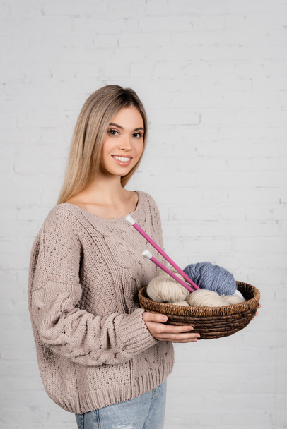 Jeune femme souriant à la caméra tout en tenant le panier avec des aiguilles à tricoter et du fil sur fond blanc - Photo, image