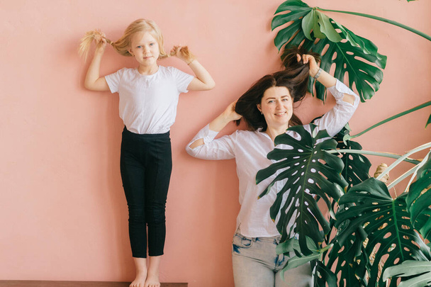 Life style female family portrait of happy mother and smiling daughter make same poses on pink wall with palm tree branches home background. Cheerful mom with her blonde daughter posing identically - Fotoğraf, Görsel