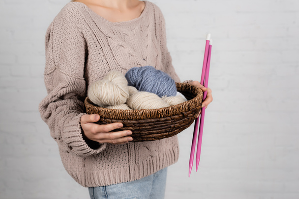Vista recortada de la mujer en la cesta de suéter con hilos de lana y agujas de punto sobre fondo blanco - Foto, Imagen
