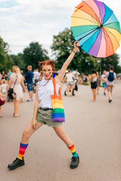 fashion girl with lgbt gay pride flag on her face, colorful umbrella, bag and mismatched socks posing on street with people on background. - Foto, imagen