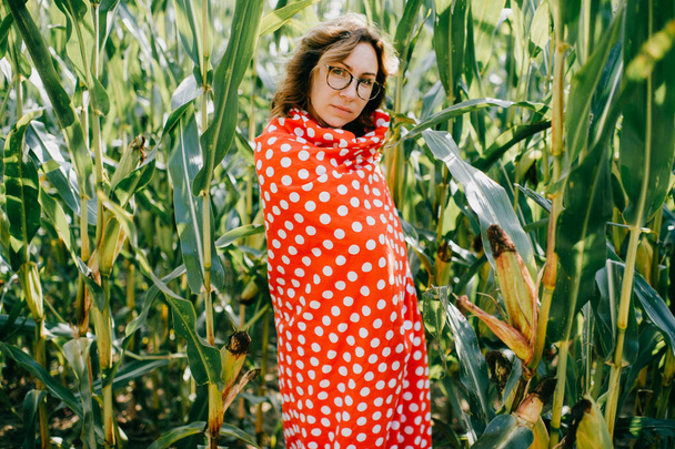 Beautiful caucasian female with short dark wavy hair in glasses wrapped in red and white coverlet enjoys of fresh breathe in cornfield. - Foto, Imagem