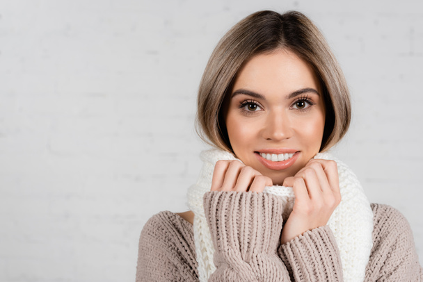 Young woman in sweater smiling at camera while holding scarf on white background - 写真・画像