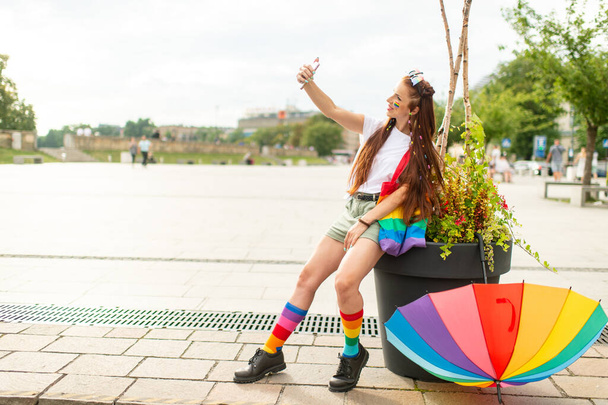 Fashionable lesbian in colorful sunglasses with lgbt rainbow on her face taking selfie with smartpthone at city square.  - Photo, Image