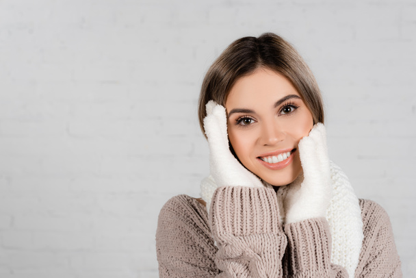 Young woman in sweater, knitted scarf and gloves with hands near face smiling at camera on white background - Фото, зображення