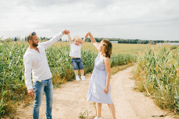Young beautiful mum in light blue dress, strong caucasian dad with short dark hair in white shirt and blue jeans playing with their cute little blond son in a cornfield in summer. - Valokuva, kuva