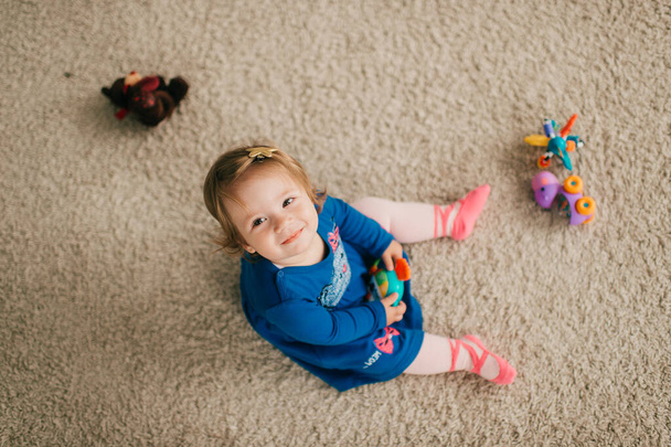 Little cute caucasian girl in attractive dress sits on the floor and carpet, plays with her various toys and looks up - Foto, immagini