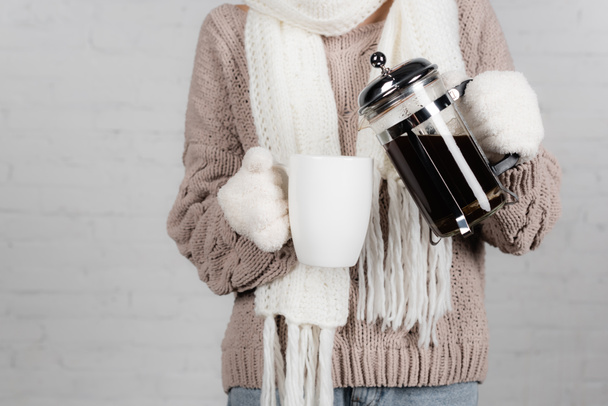Cropped view of woman in knitted sweater, scarf and warm gloves pouring coffee from pot in cup on white background - Photo, Image