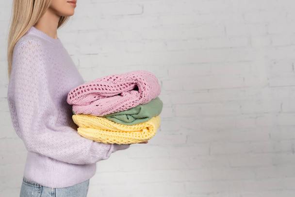 Cropped view of woman holding colorful warm sweaters near white brick wall - Фото, зображення