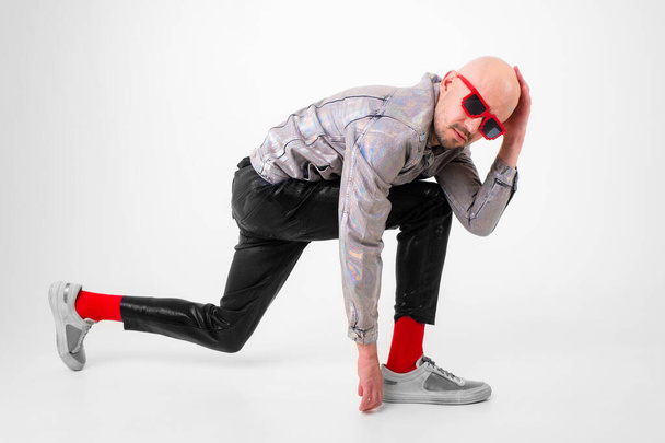 Beautiful caucasian man without hair in grey shirt, black leather trousers, black sunglasses, red socks and grey trainers poses for the camera isolated on white background. - Fotografie, Obrázek
