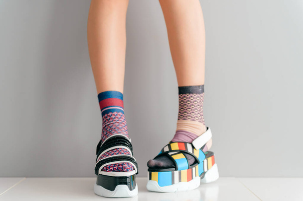 Beautiful female legs in mismatched trendy socks standing in two different fashionable high wedge leather sandals on white surface. Odd disargonized  young girl wearing high sole summer stylish shoes - 写真・画像