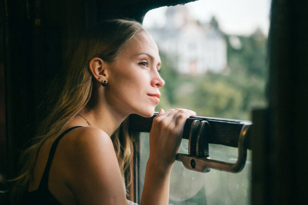 Mood atmospheric lifestyle portrait of young beautiful blonde hair girl looking out of window from riding train. Pretty teen enjoying beauty of nature from moving train car in summer. Travel concept. - Photo, image