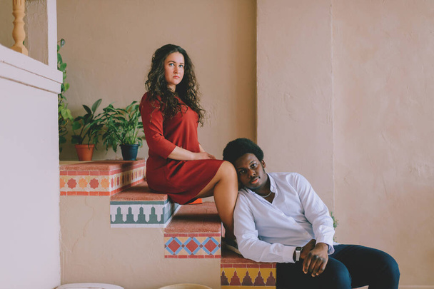 Interracial loving couple lifestyle indoor portrait. Dark skinned young african man siiting on stairs with caucasian girlfriend. Black guy put his head on woman knees. International family portrait - Фото, изображение