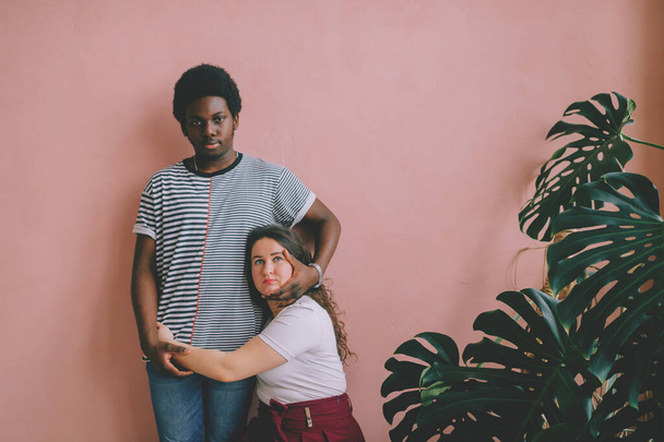 Hugging interracial couple lifestyle indoor portrait on pink wall background. Dark skinned nigerian man in striped shirt embracing caucasian girlfriend in red dress at home. International friendship - 写真・画像
