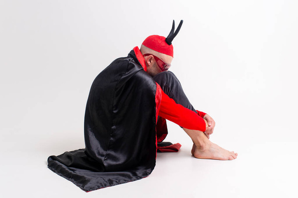 Srange unhappy man in dracula  masquerade costume and devil horns sitting in depression and bad feelings over white background and hiding his face in the knees. - Zdjęcie, obraz