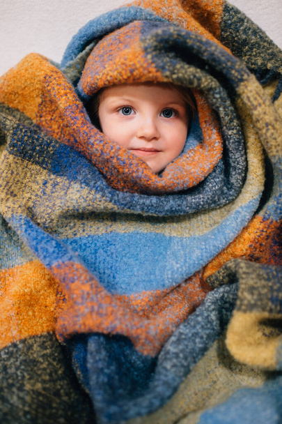 Pretty caucasian little girl with fair hair in blue dress sits in baby room wrapped herself in a colourful coverlet and smiles - Fotografie, Obrázek
