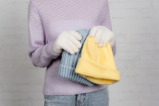 Cropped view of woman in sweater and warm gloves holding colorful hats on white background - Photo, Image