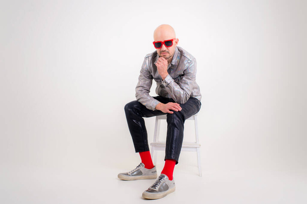 Funny caucasian man without hair in grey shirt, black leather trousers, black sunglasses, red socks and grey trainers sits on a chair isolated on white background. - Фото, изображение