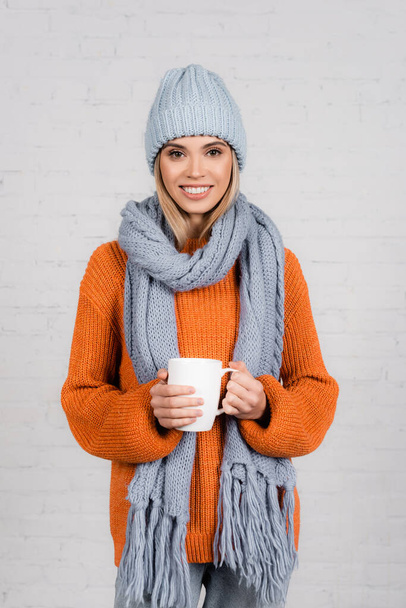 Young woman in knitted sweater, hat and scarf holding cup and smiling on white background - Foto, afbeelding